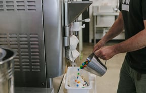 A man pouring M&M's into the gelato tub while gelato is falling out of the machine.