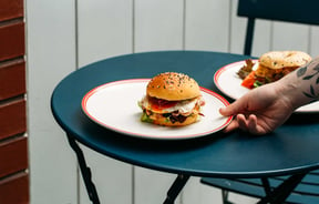 A hand putting a burger on a plate on to a blue table.