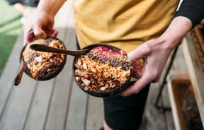 A staff member holds two bowls, made from coconut husks, of acai topped with crunchy coconut, cacao nibs and buckwheat groats.