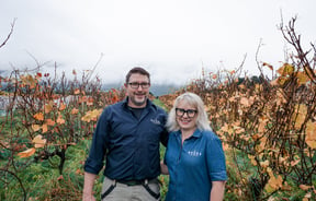 A man and woman smiling to camera in a vineyard on an autumn day.
