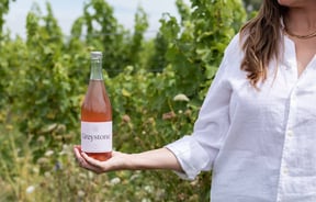 A woman in a white shirt holds a bottle of Greystone Rose Pet Nat against the vineyard's green backdrop at Greystone Wines.