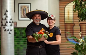 A man and woman holding a bowl of fresh vegetables smiling to camera.
