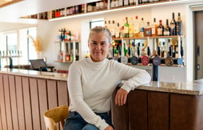 A blonde woman sitting at a bar smiling to camera.