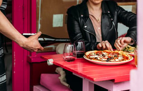 Wine being poured into a glass next to a woman pulling at a pizza.