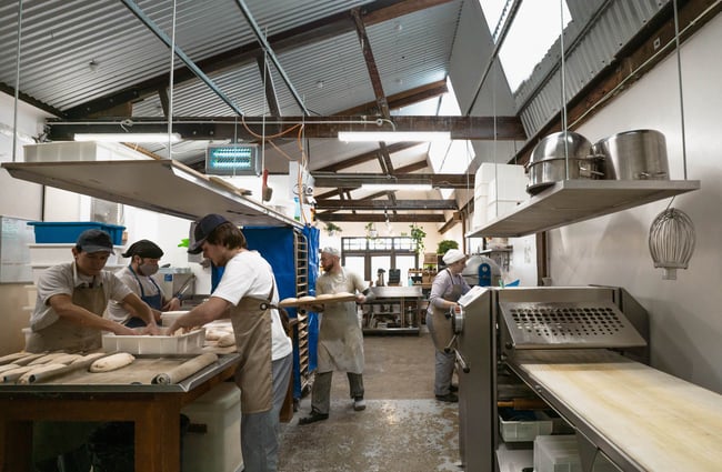 Bakers, dressed in white with stone brown aprons, working in the kitchen at Bellbird Bakery.