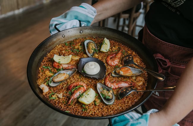 A close up of a pan of paella being carried by a staff member.