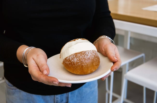 Hands holding a plate with a cream bun on top.