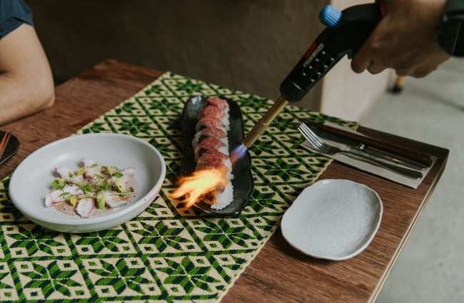 Fire being used to cook the top of sushi rolls on a dining table.