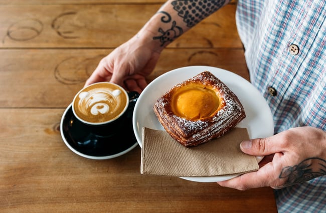 Close up image of a staff member holding an apricot Danish and a barista coffee topped with a bear face drawn with the steamed milk.