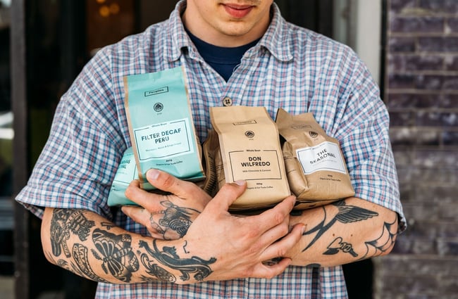 Close up of a man in a short-sleeve checkered shirt with tattooed arms holding four bags of Peoples Coffee beans to his chest.