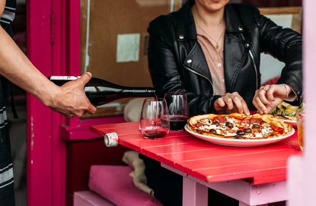 A staff member pouring wine into a glass whilst a customer pulls at a pizza.