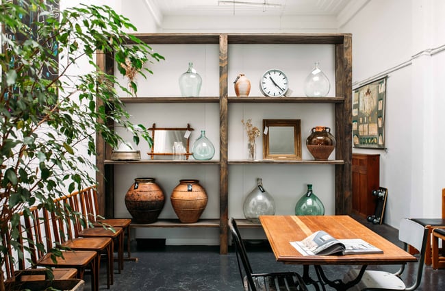 Large vases, mirrors and clocks on cabinetry, sitting alongside wooden chairs and a table.