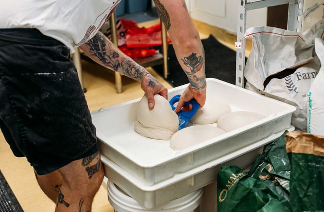 A tattooed staff member scooping up a ball of dough ready to be made into a pizza.