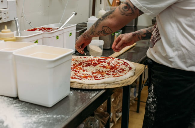 A tattooed man sprinkling cheese on an uncooked pizza.