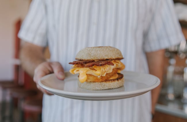 A hand holding a bacon and egg bun on a plate.