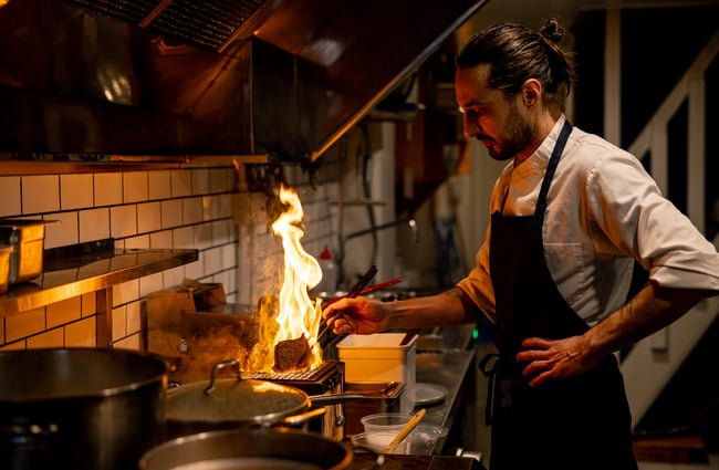 A male chef working with fire inside a restaurant kitchen.