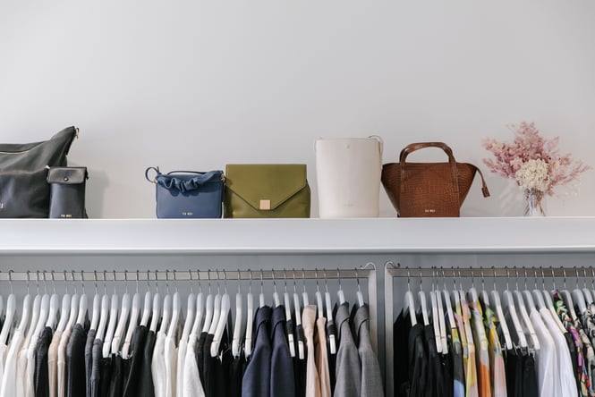 Yu Mei handbags sit on a shelf above a rack of hanging clothes.