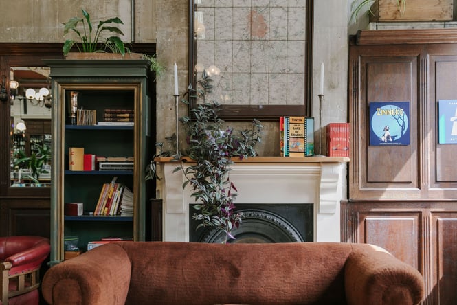A brown suede couch in front of an old fire place decorated with books and plants.