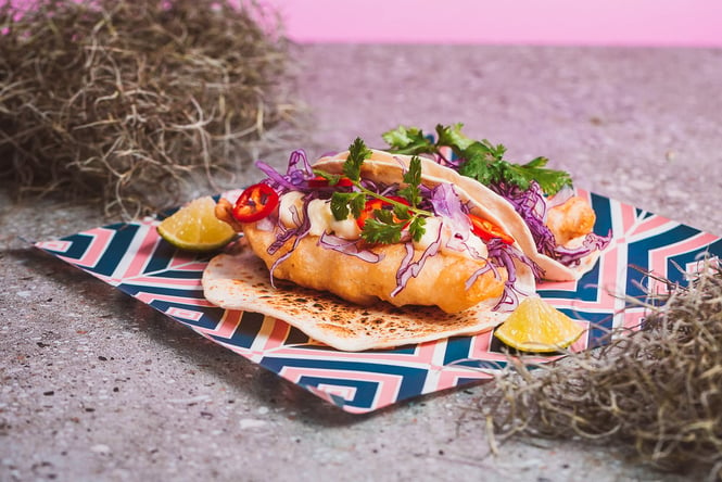 A close up of a fish taco on a colourful piece of paper.
