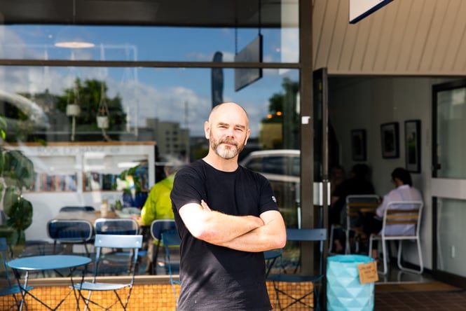 A man with arms crossed looking to camera outside a cafe.