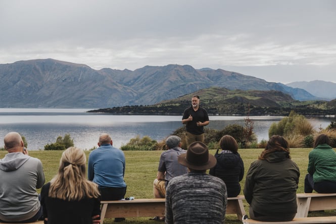 A bearded man presenting to a group of people outside in front of a lake.