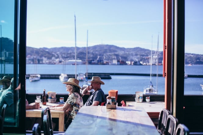 People dining on outdoor deck next to the ocean on a sunny Wellington day.