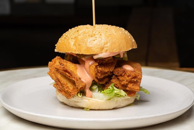 A fried chicken burger on a white plate with a wooden skewer holding it together.