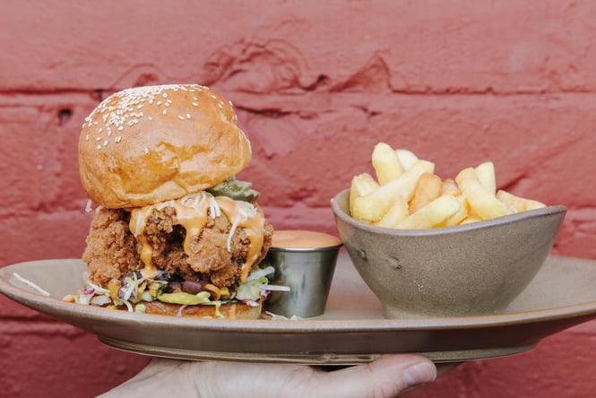 Someone holding a plate in front of a red wall with a fried chicken burger and a bowl of fries.