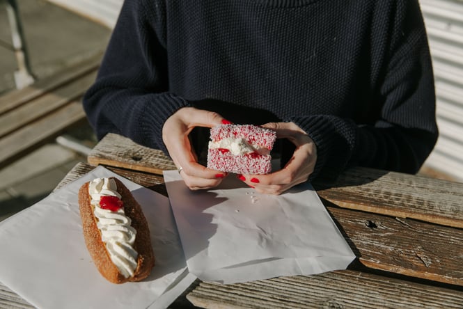 Two hands holding a pink lamington next to a cream bun at an outside table.