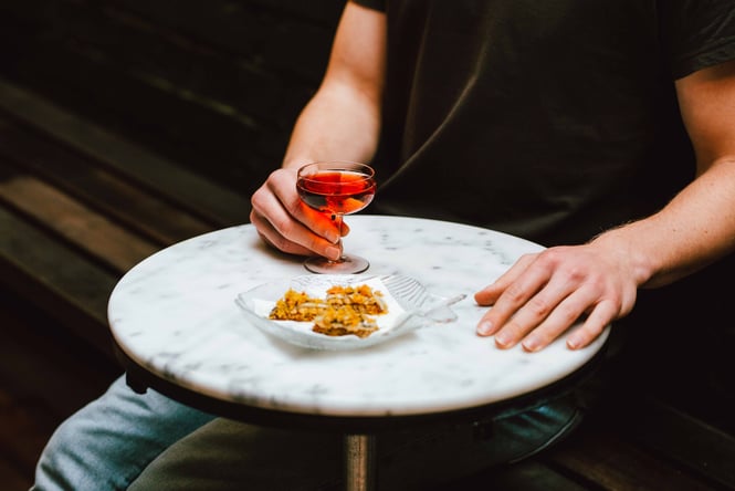 A hand holding a glass of pink wine with a small plate of snacks on the table.