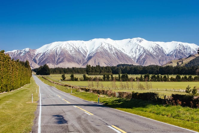 A country road on a sunny day with snowy mountains in the background.