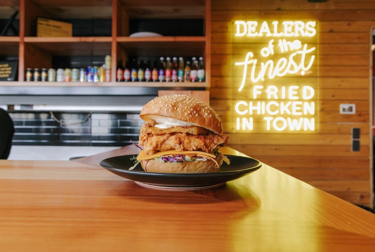 A Chicken burger on a black plate sitting on a wooden bench.