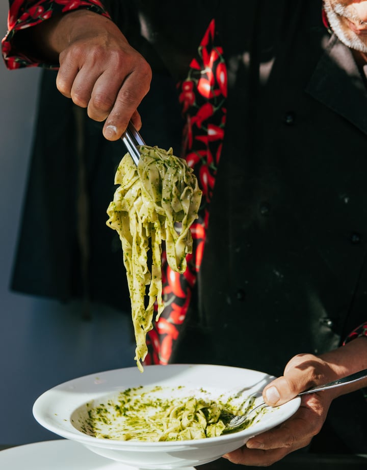 A chef serving a plate of spinach pasta on a plate in the sunlight.