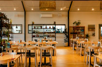 Contemporary and plant-filled interior of Off The Track restaurant with the light wooden dining tables and a chef cooking through the open kitchen window