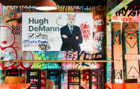A bar leaner and bar stools up against a graffiti covered wall.
