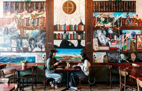 Two diners sitting at a table eating with lots of decorative  artwork behind them.