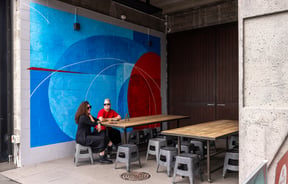 Two people sitting at table in front of bright mural at CoCA Gallery in Christchurch