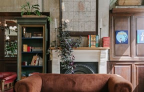 A brown suede couch in front of an old fire place decorated with books and plants.