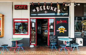The exterior of Deluxe with its red-framed door and window and round tables and chairs set out on the brick-paved footpath.
