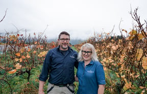 A man and woman smiling to camera in a vineyard on an autumn day.
