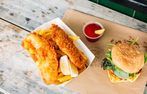 A flatlay of fish and chips and burger with a little pottle of sauce on an outdoor table.