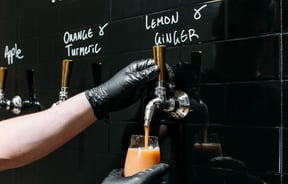 A worker in black gloves pouring lemon and ginger kombucha from the tap.