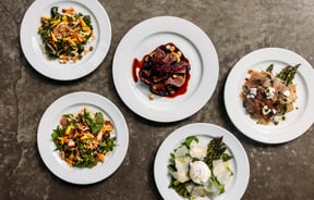 Flat lay of five beautiful plates of food including thai salads, prosciutto, venison and asparagus