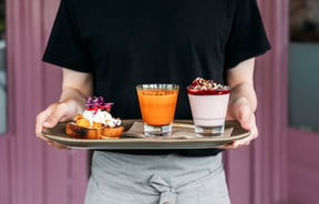 Waiter holding breakfast board with yogurt and fruit, an orange and carrot juice, and a piece of toast with a poached egg and pickles on top