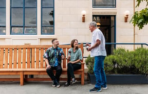 Waiter serving wine to two guests sitting on bench chair outside wine bar