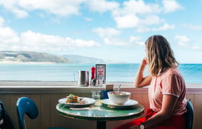 Customer sitting at an outdoor table at Maranui Cafe with the sea behind her.