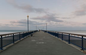A couple walking along New Brighton pier in the evening with pinky skies and calm waters in the background