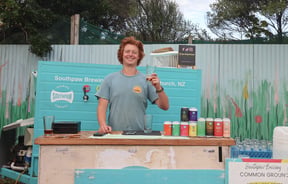 A man holding a beer and smiling to camera outside.