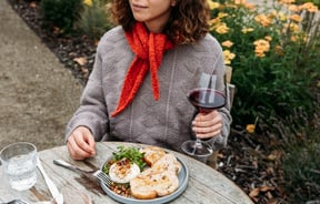 Woman sitting at an outdoor table with a glass of wine in her hand and dish of food in front of her on the table.