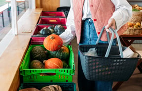 A person holding a shopping basket picks an orange pumpkin out of a plastic crate.